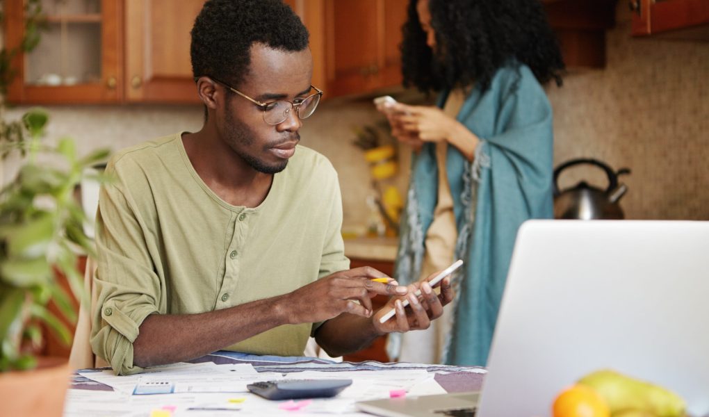 Dark-skinned man in spectacles doing finances, using cell phone, calculator and laptop computer, making call to utility services. Female with Afro hairstyle standing in background with mobile