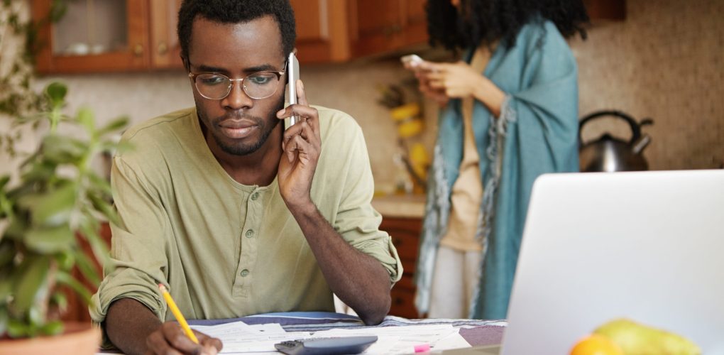 Serious African man having phone conversation with bank asking to extend loan term for paying out mortgage, holding pencil in the other hand, making notes in documents, lying on table in front of him