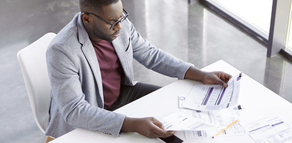 Top view of serious confident young dark skinend male economist, uses paper documents, checks monthly report, has confident expression, sits at his cabinet. People, job and business concept.